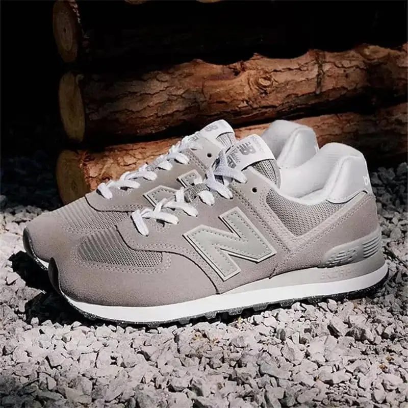 A classic New Balance 574 sneaker featuring a combination of gray suede and mesh, white laces, and a prominent blue-outlined “N” logo. The shoe has a cream midsole with a black outsole, offering a clean, versatile look perfect for everyday casual wear.